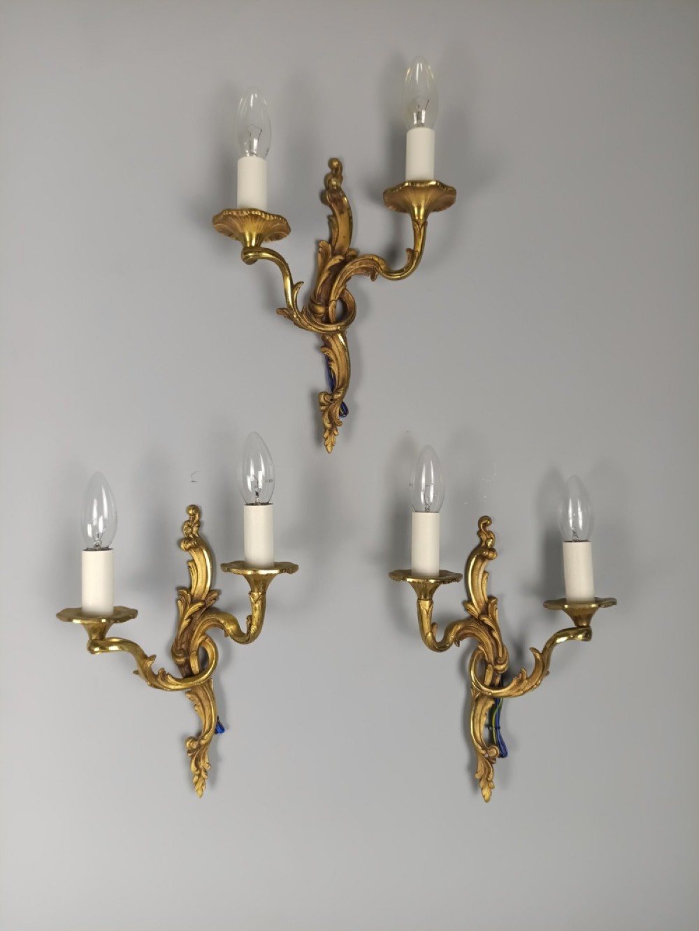 Set of three French Rococo style gilt bronze wall sconces featuring twin sweeping arms, foliate scroll backplates, and candle-effect fittings.