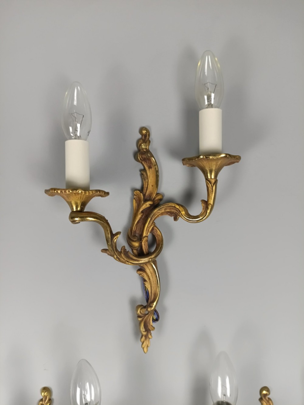 Set of three French Rococo style gilt bronze wall sconces featuring twin sweeping arms, foliate scroll backplates, and candle-effect fittings. View V