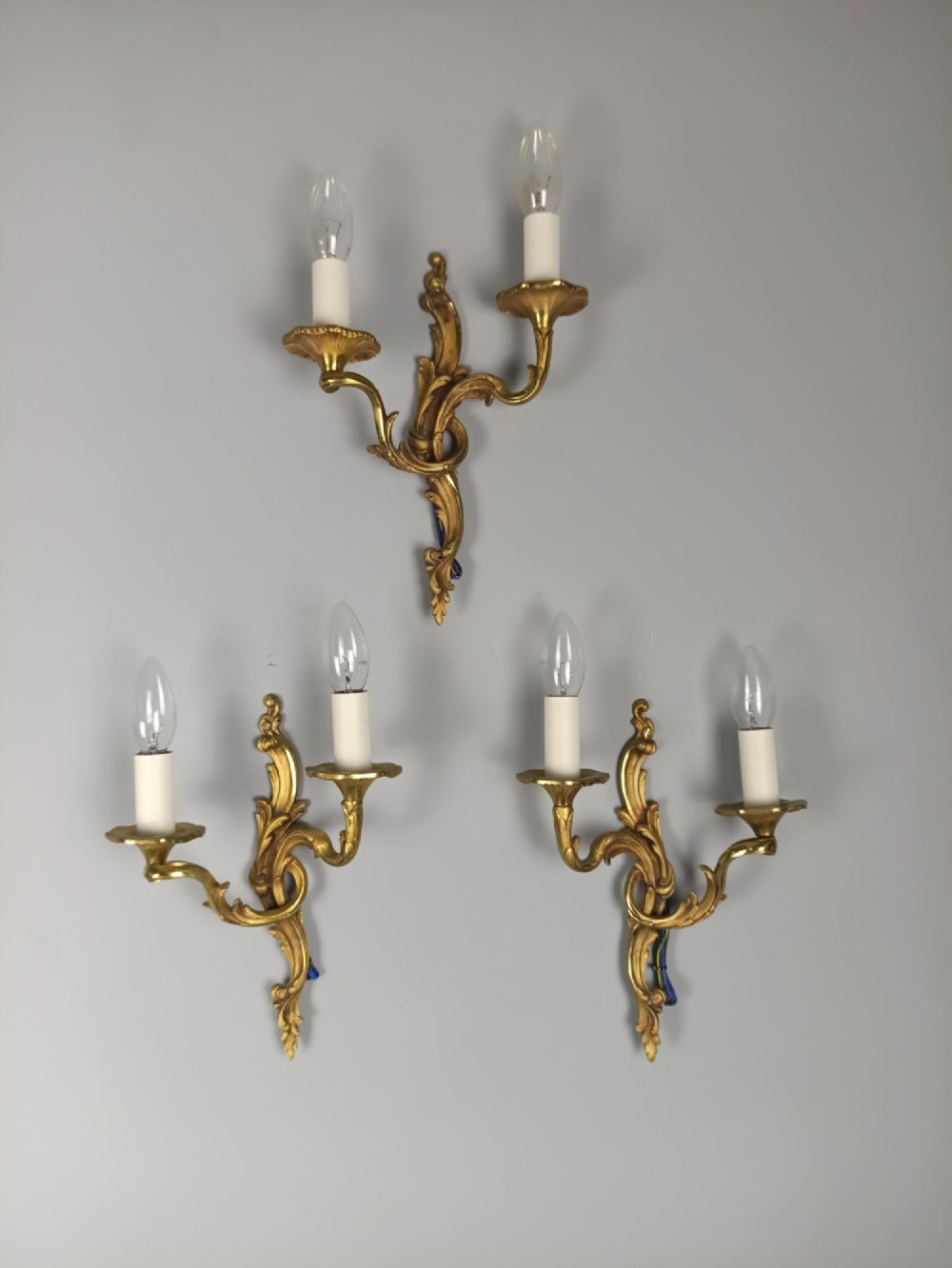 Set of three French Rococo style gilt bronze wall sconces featuring twin sweeping arms, foliate scroll backplates, and candle-effect fittings. View VI