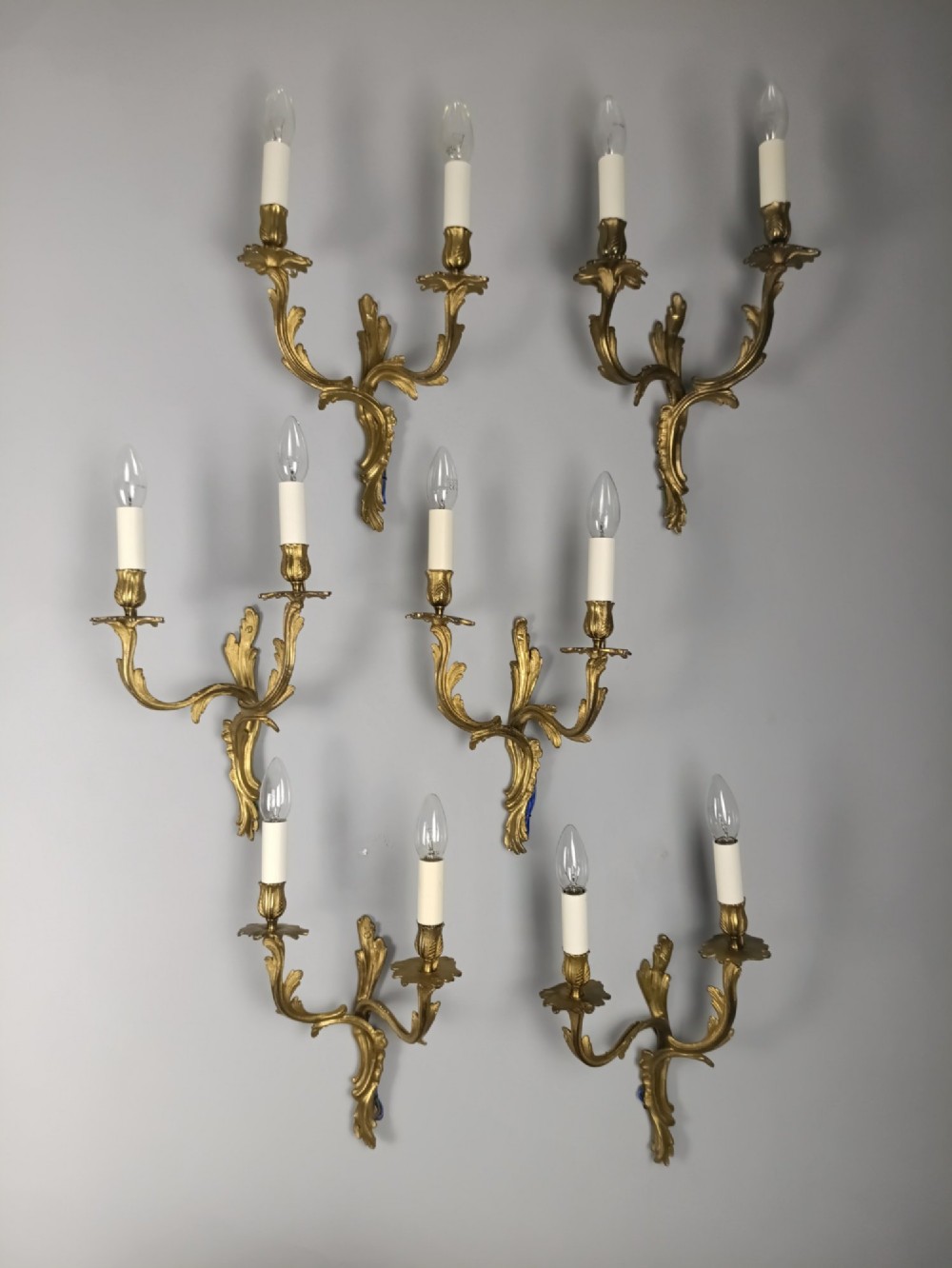 A set of six French Rococo style twin-arm gilded brass wall lights featuring acanthus leaf scrollwork and white candle sleeves.