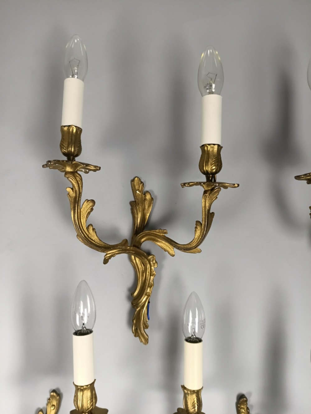 A set of six French Rococo style twin-arm gilded brass wall lights featuring acanthus leaf scrollwork and white candle sleeves. View III