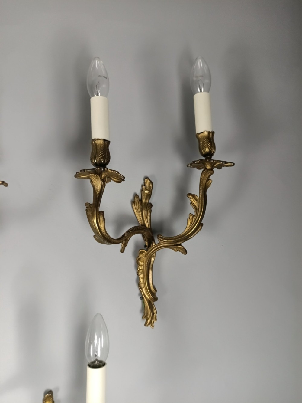 A set of six French Rococo style twin-arm gilded brass wall lights featuring acanthus leaf scrollwork and white candle sleeves. View IV