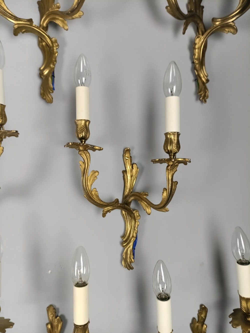 A set of six French Rococo style twin-arm gilded brass wall lights featuring acanthus leaf scrollwork and white candle sleeves. View V