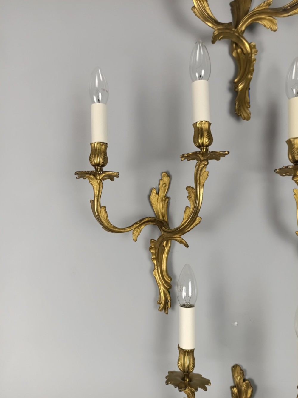 A set of six French Rococo style twin-arm gilded brass wall lights featuring acanthus leaf scrollwork and white candle sleeves. View VI