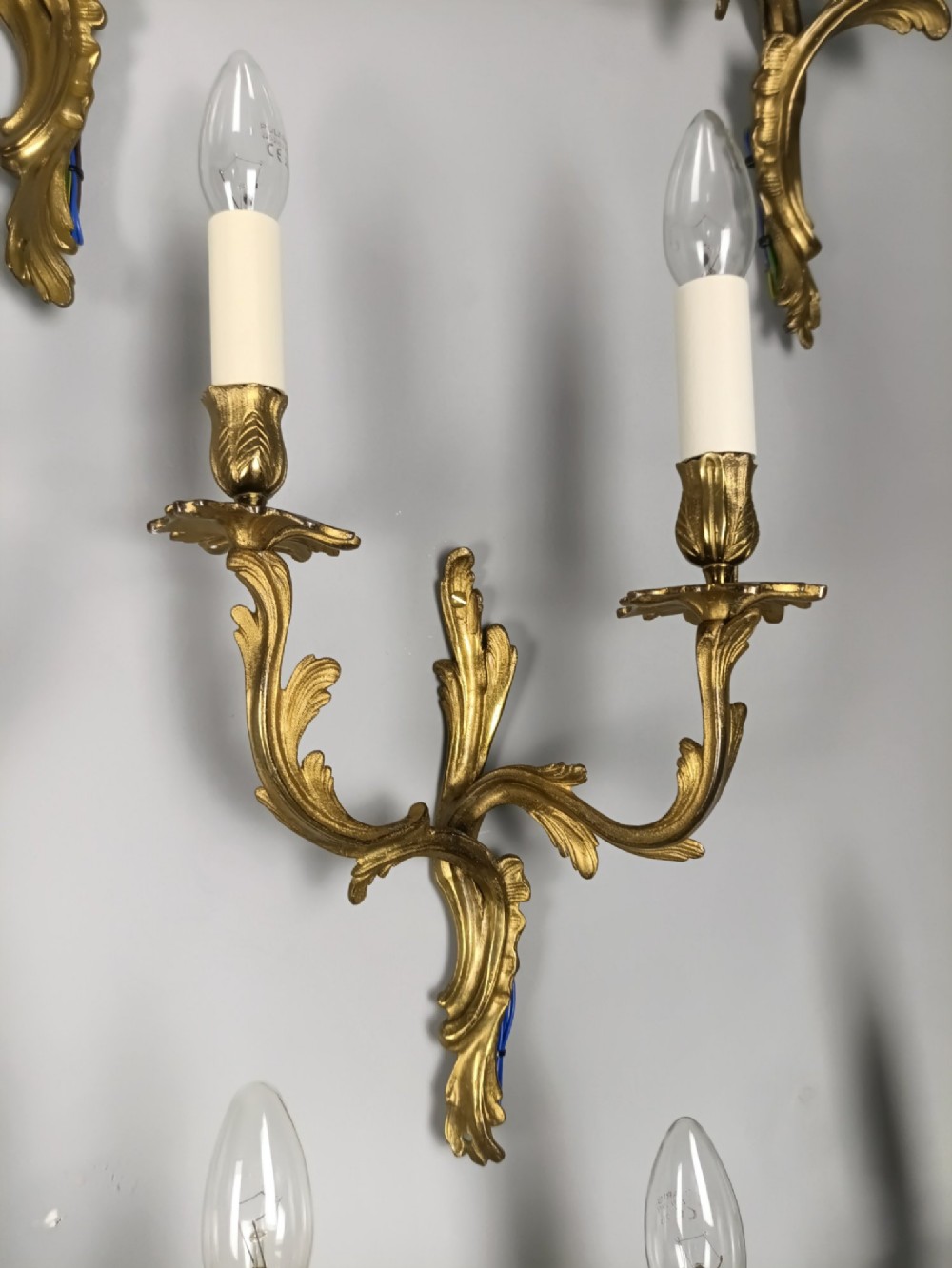 A set of six French Rococo style twin-arm gilded brass wall lights featuring acanthus leaf scrollwork and white candle sleeves. View IX