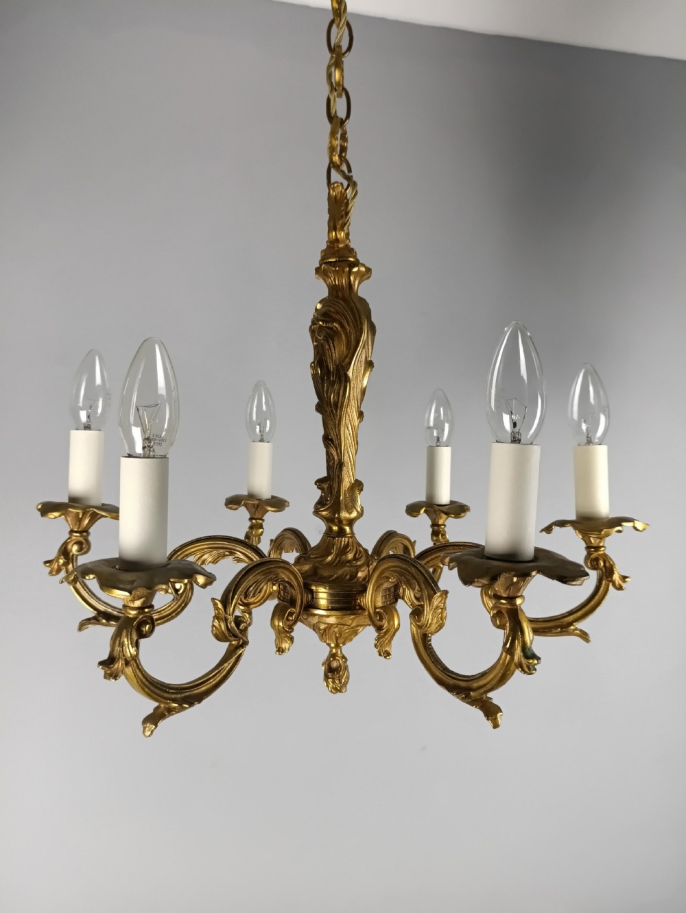 A French six-light gilt bronze chandelier featuring scrolling acanthus leaf arms, candle-style bulb sleeves, and an ornamental central stem suspended from a chain.