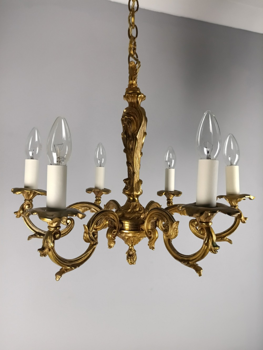 A French six-light gilt bronze chandelier featuring scrolling acanthus leaf arms, candle-style bulb sleeves, and an ornamental central stem suspended from a chain. View II