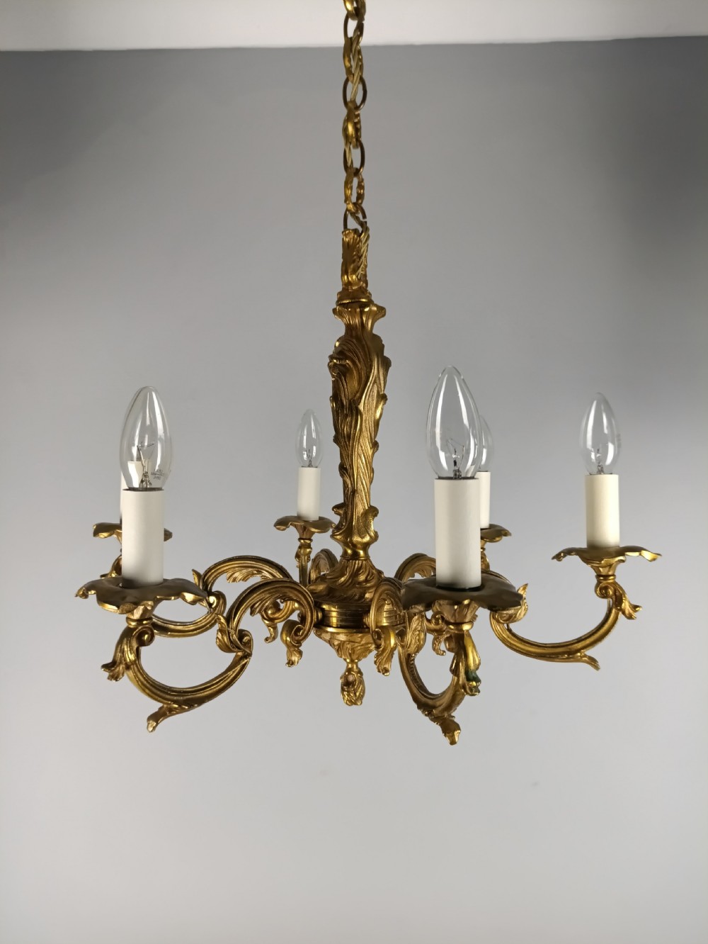 A French six-light gilt bronze chandelier featuring scrolling acanthus leaf arms, candle-style bulb sleeves, and an ornamental central stem suspended from a chain. View IV
