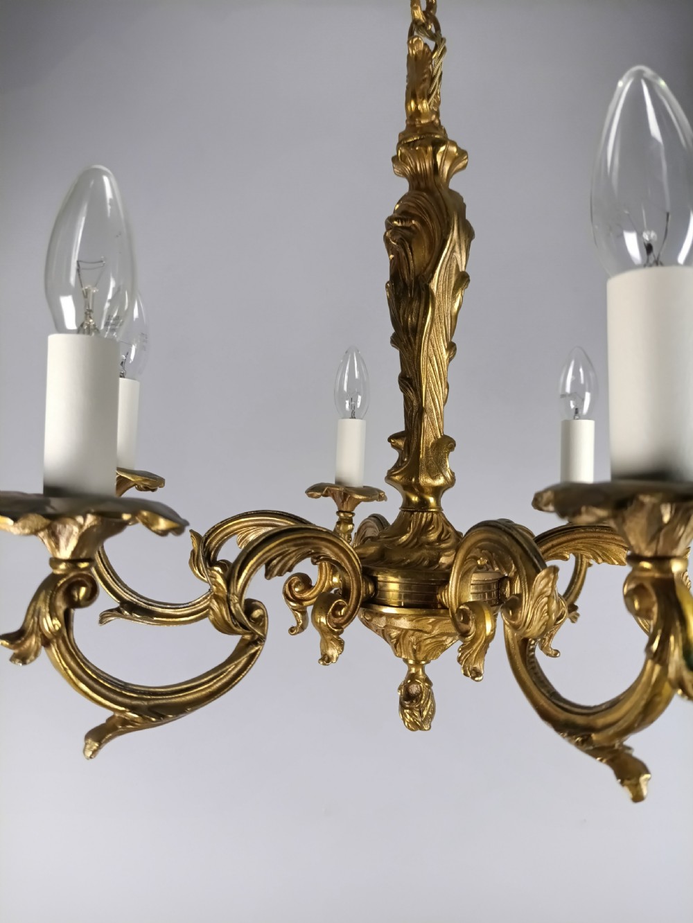 A French six-light gilt bronze chandelier featuring scrolling acanthus leaf arms, candle-style bulb sleeves, and an ornamental central stem suspended from a chain. View V