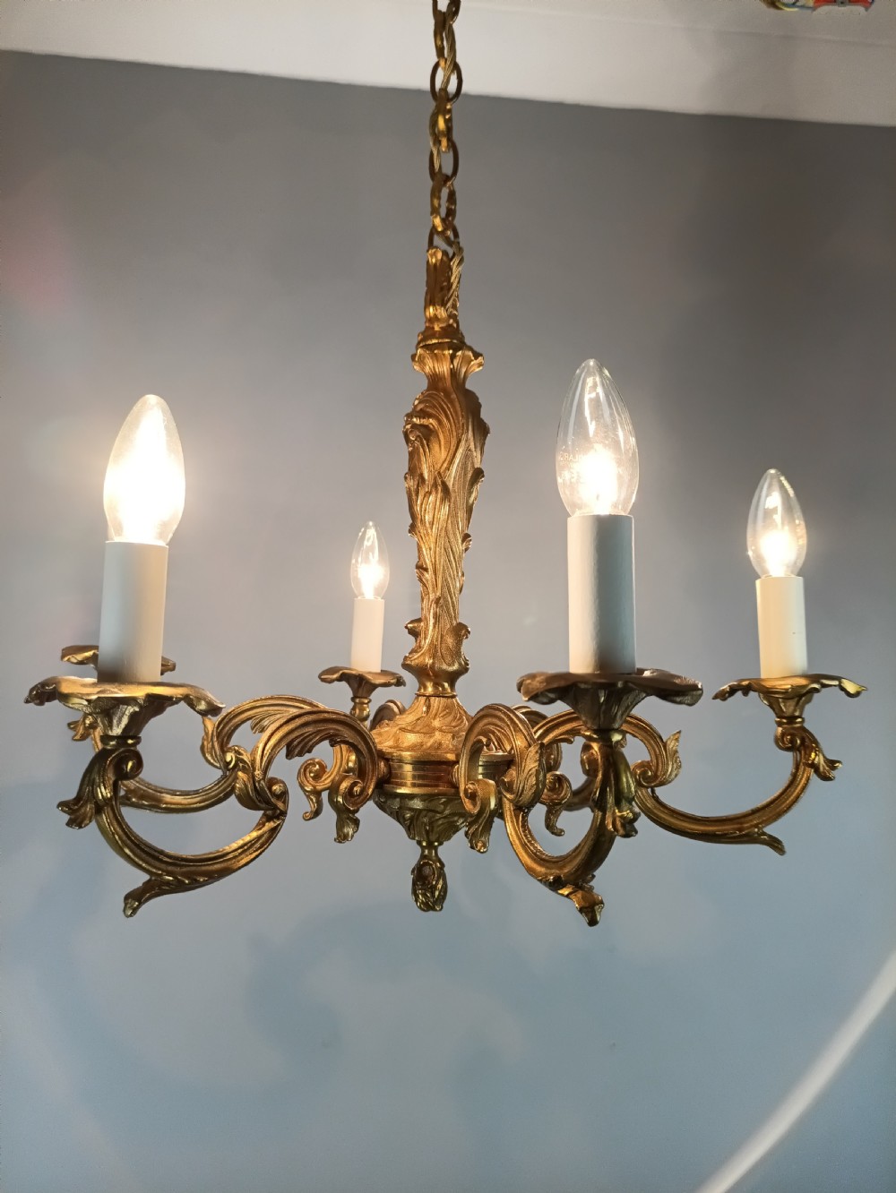 A French six-light gilt bronze chandelier featuring scrolling acanthus leaf arms, candle-style bulb sleeves, and an ornamental central stem suspended from a chain. View X