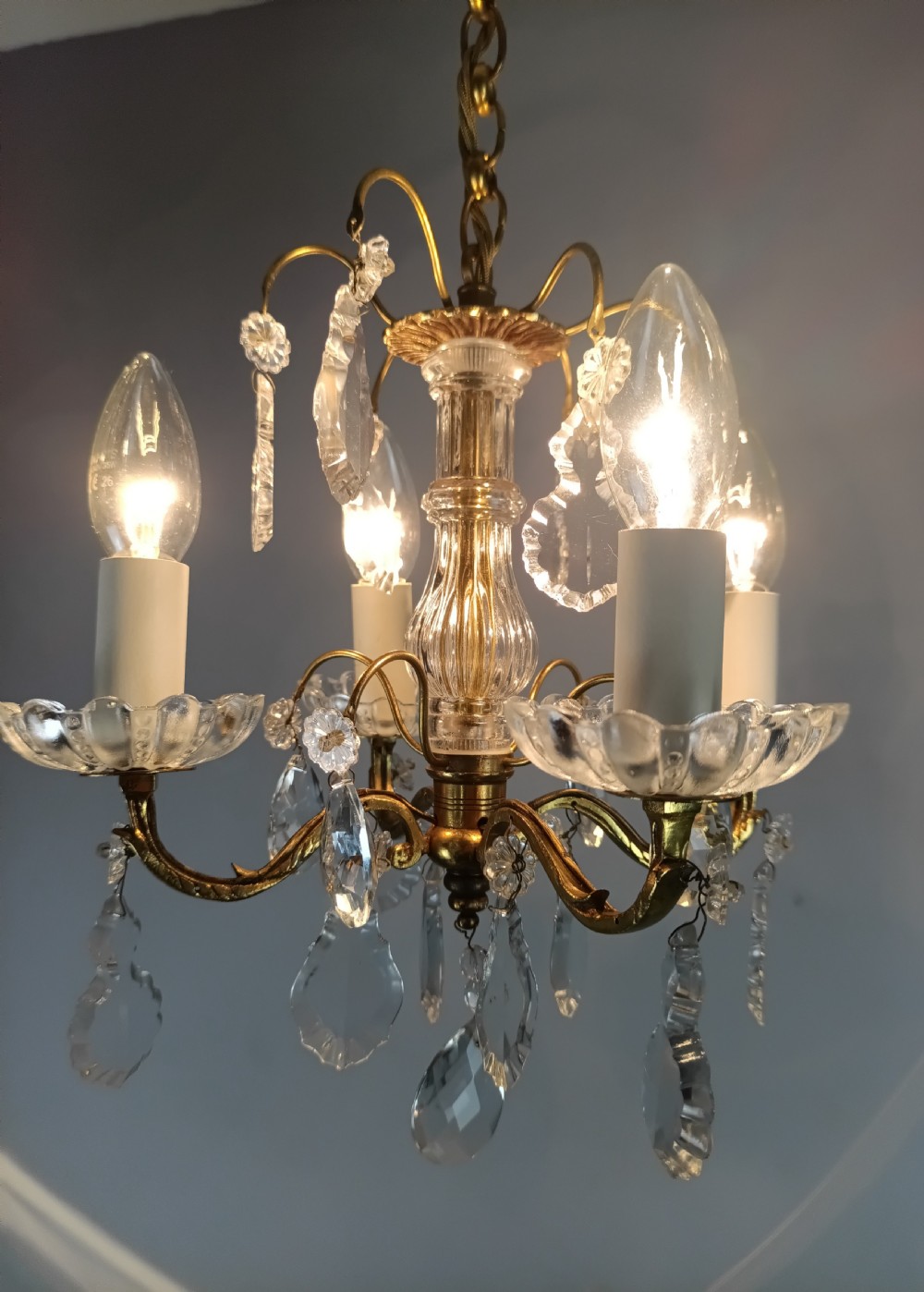 A four-arm brass mini chandelier featuring clear glass droplets, floral bobeches, and a central glass stem column. View X