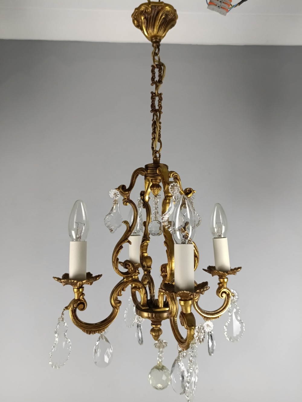 A four-arm French gilt metal chandelier decorated with scrolling arms, candle-style light sockets, and hanging faceted cut crystal drops. View IV
