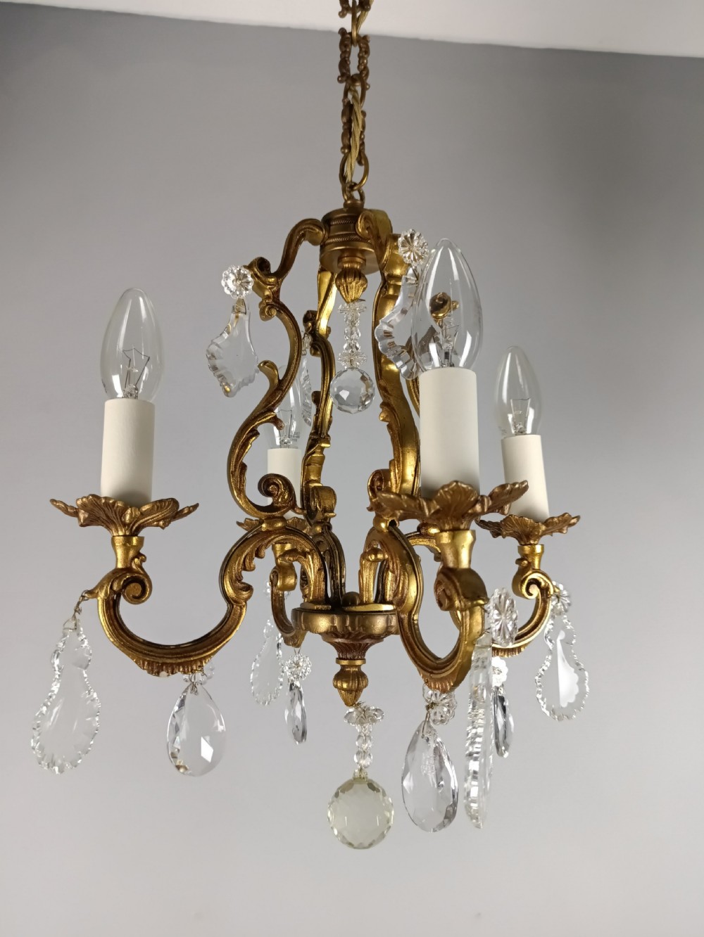 A four-arm French gilt metal chandelier decorated with scrolling arms, candle-style light sockets, and hanging faceted cut crystal drops. View V