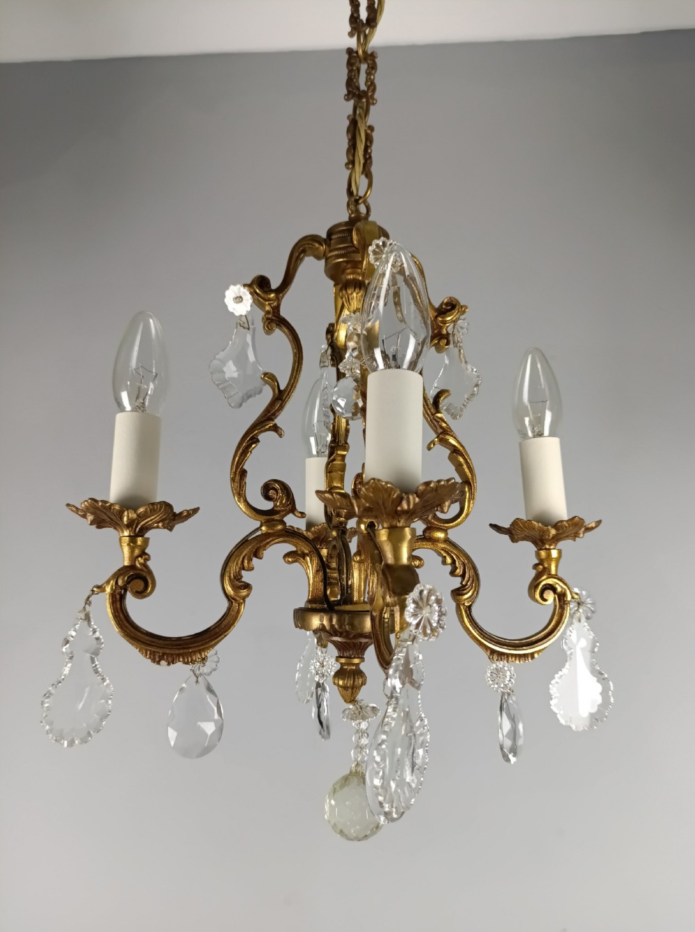 A four-arm French gilt metal chandelier decorated with scrolling arms, candle-style light sockets, and hanging faceted cut crystal drops. View VI