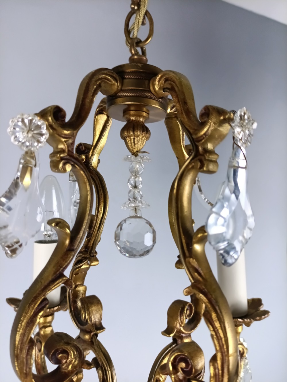 A four-arm French gilt metal chandelier decorated with scrolling arms, candle-style light sockets, and hanging faceted cut crystal drops. View VII