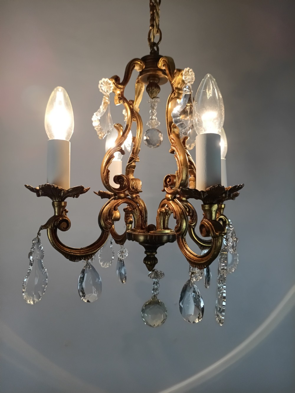 A four-arm French gilt metal chandelier decorated with scrolling arms, candle-style light sockets, and hanging faceted cut crystal drops. View X