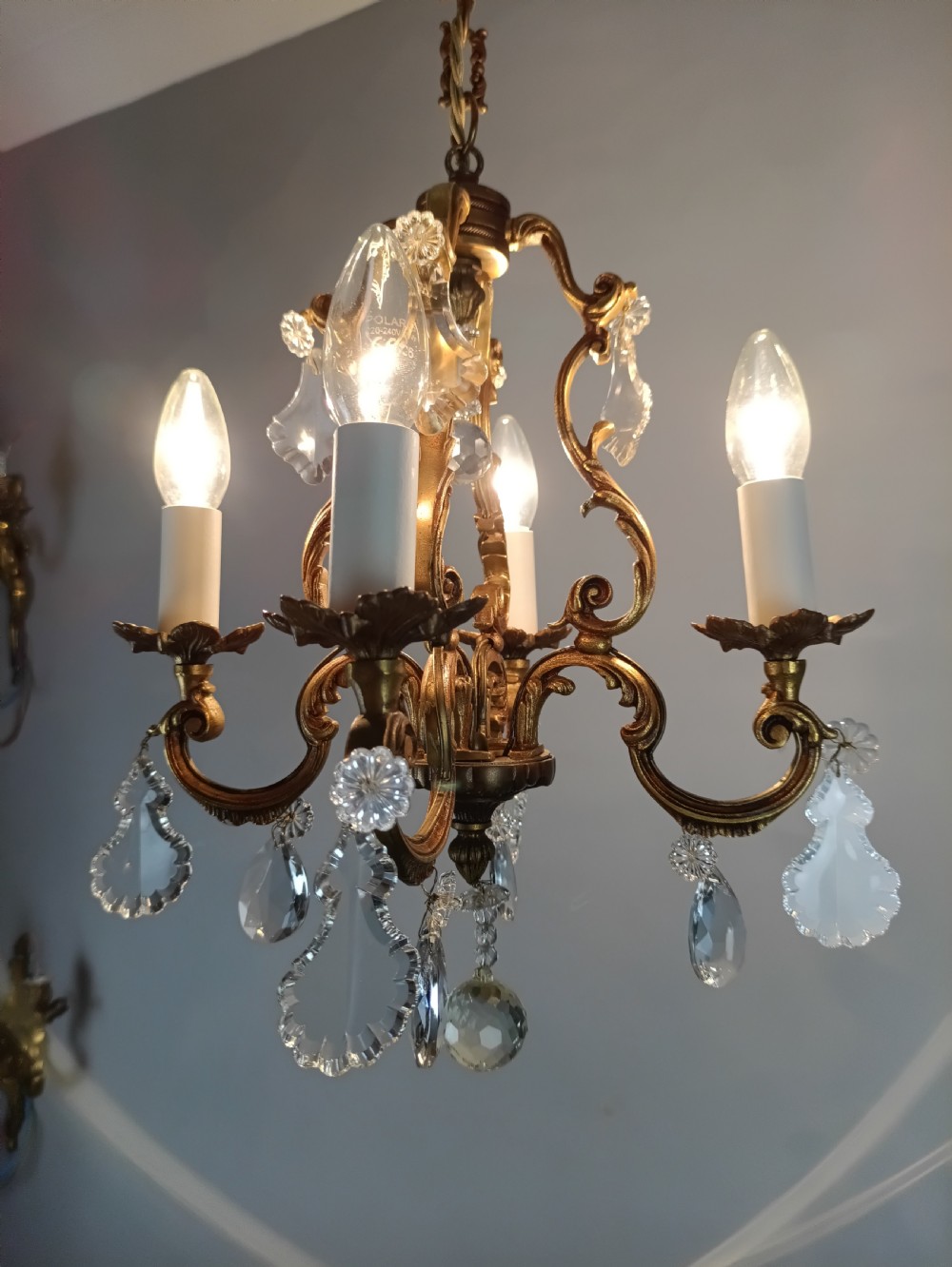 A four-arm French gilt metal chandelier decorated with scrolling arms, candle-style light sockets, and hanging faceted cut crystal drops. View XI