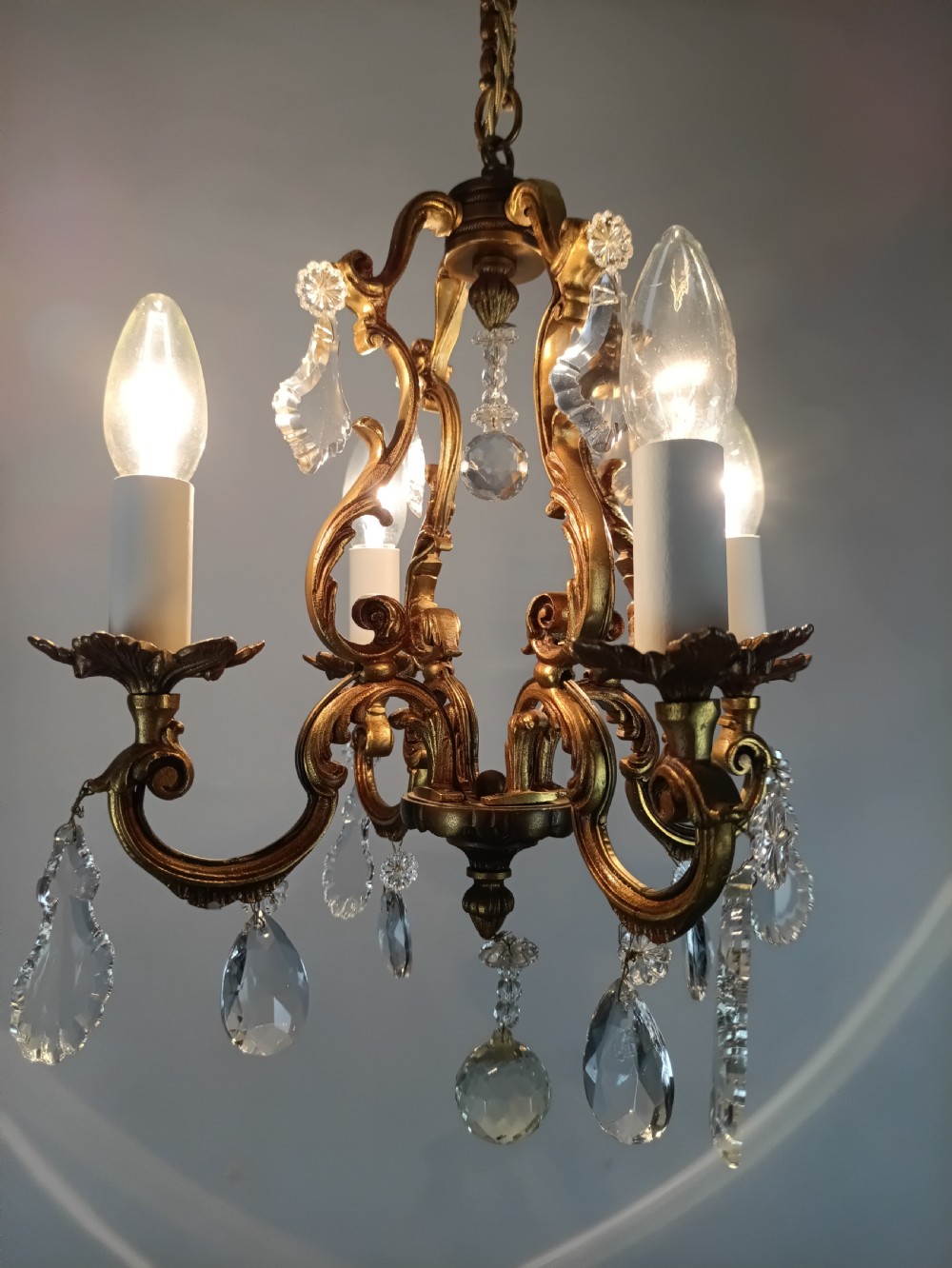 A four-arm French gilt metal chandelier decorated with scrolling arms, candle-style light sockets, and hanging faceted cut crystal drops. View XII