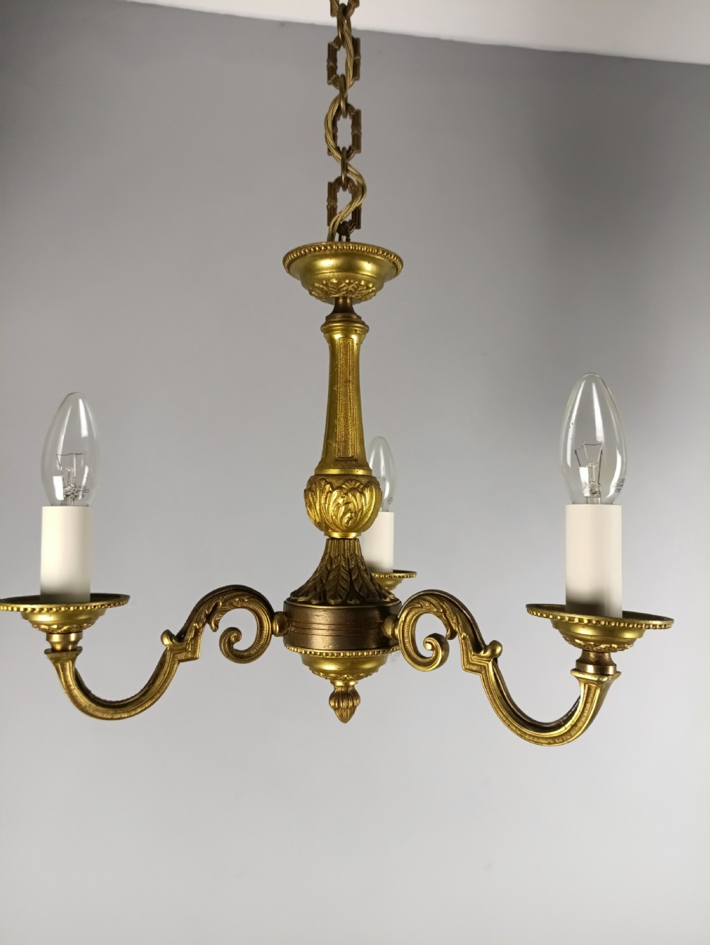 A three-arm gilt brass ceiling chandelier featuring scrolled branches, acanthus leaf decoration, and a central tapered column suspended by a chain.