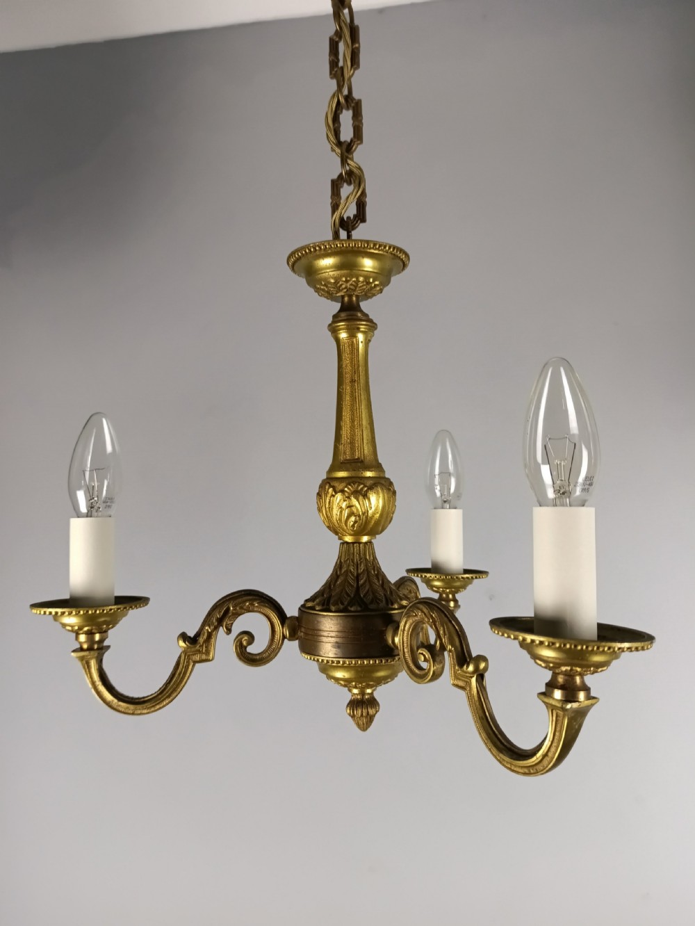 A three-arm gilt brass ceiling chandelier featuring scrolled branches, acanthus leaf decoration, and a central tapered column suspended by a chain. View IV