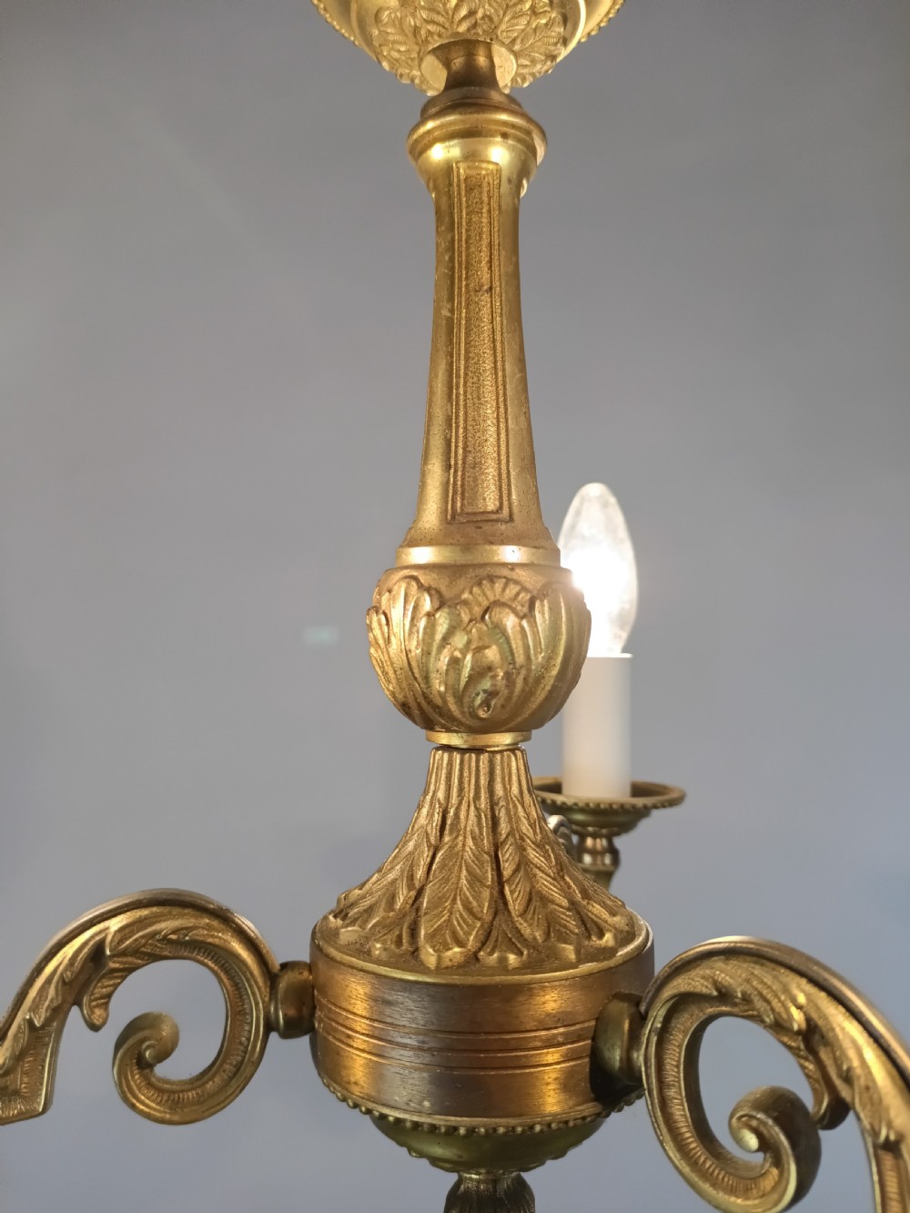 A three-arm gilt brass ceiling chandelier featuring scrolled branches, acanthus leaf decoration, and a central tapered column suspended by a chain. View X