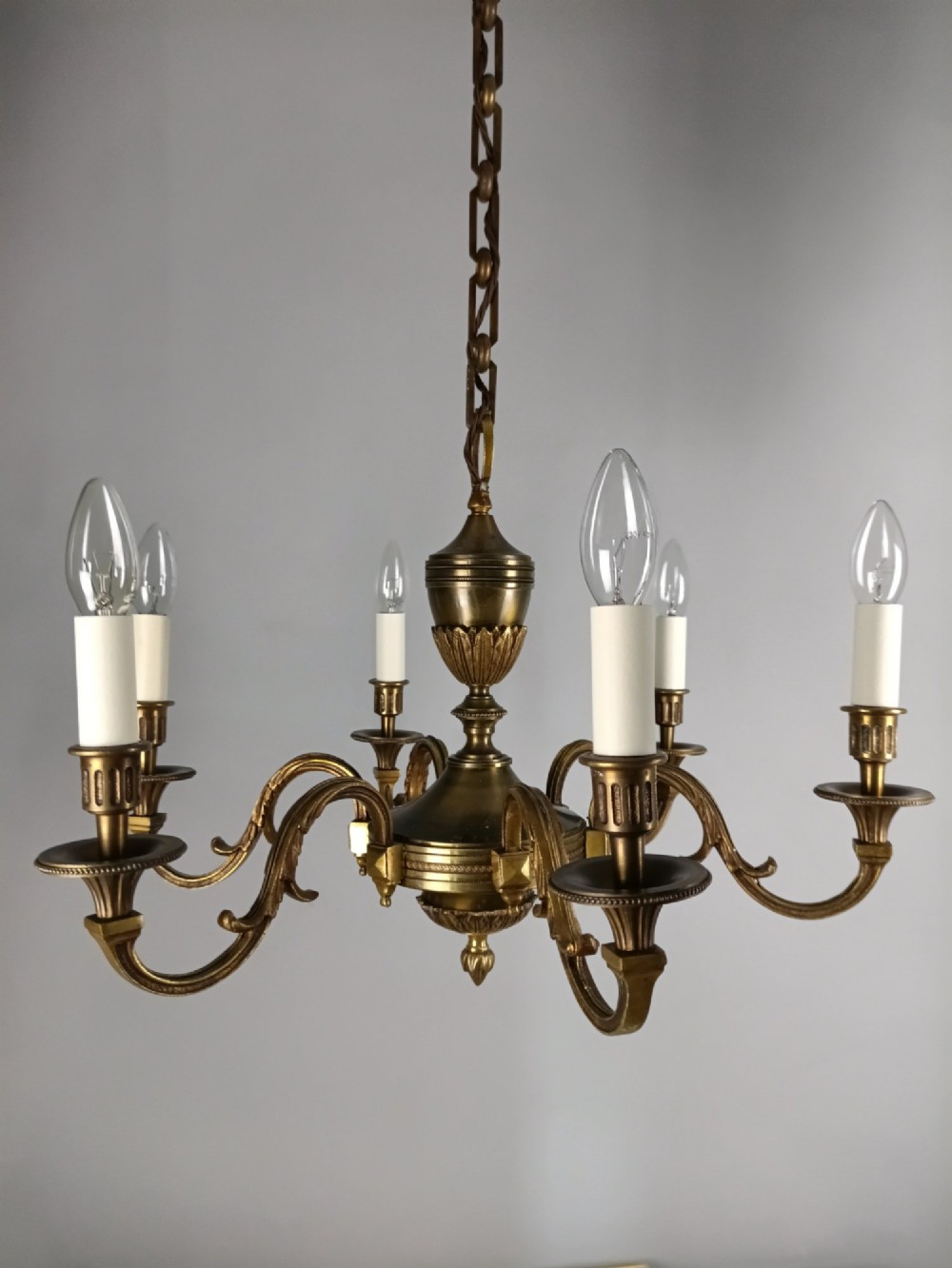 A French antique-style bronze six-arm chandelier featuring scrolling candle arms, white candle sleeves, and a central urn-shaped stem suspended from a metal chain.