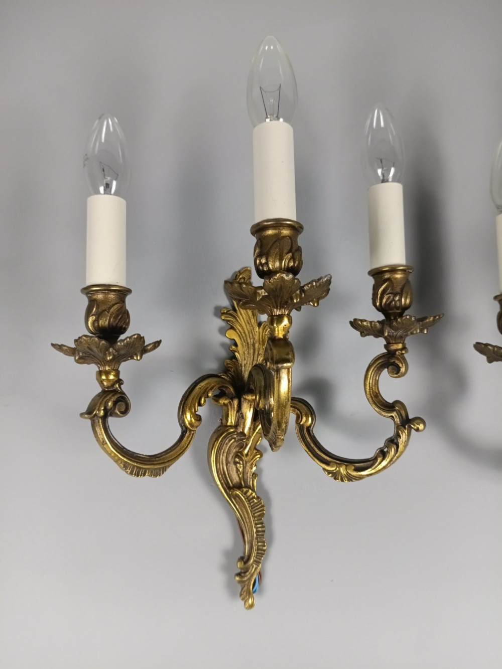 A pair of French Rococo style triple-arm gilded bronze wall lights featuring ornate foliage scrollwork and white candle sleeves. View II