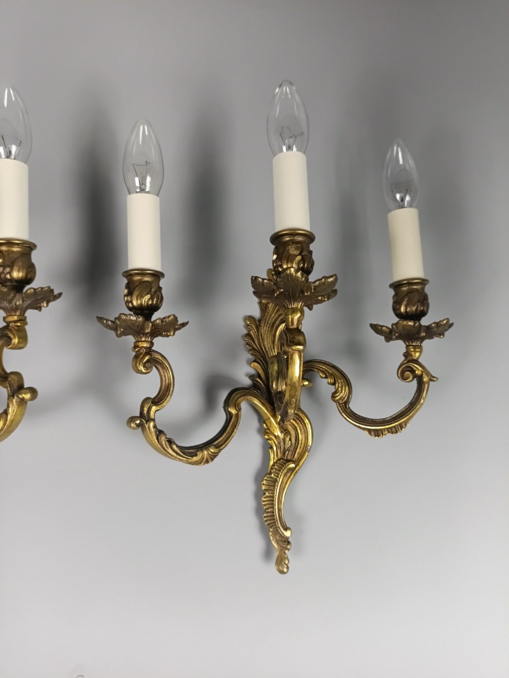 A pair of French Rococo style triple-arm gilded bronze wall lights featuring ornate foliage scrollwork and white candle sleeves. View III