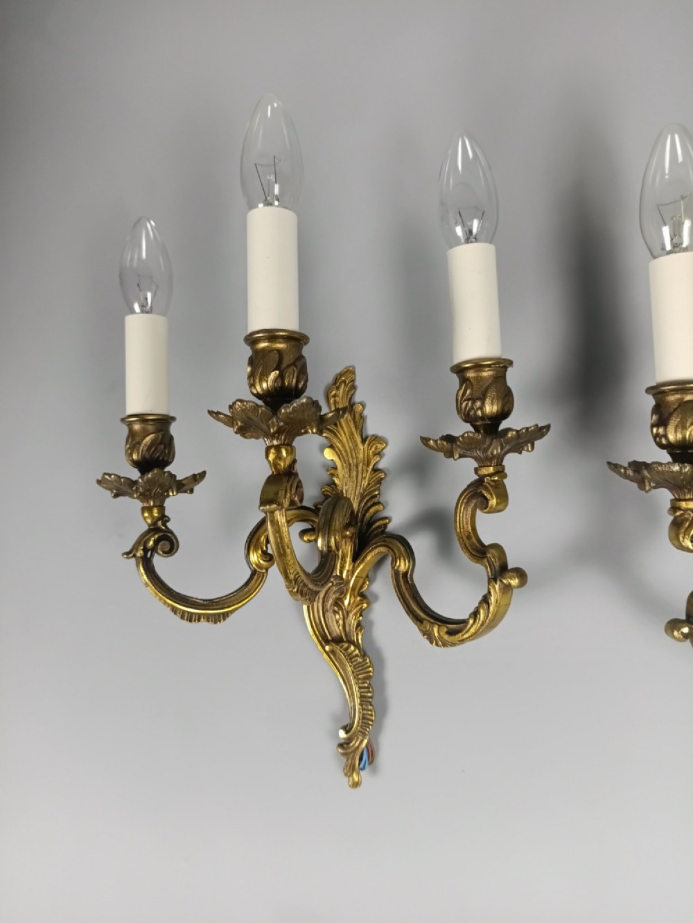 A pair of French Rococo style triple-arm gilded bronze wall lights featuring ornate foliage scrollwork and white candle sleeves. View IV