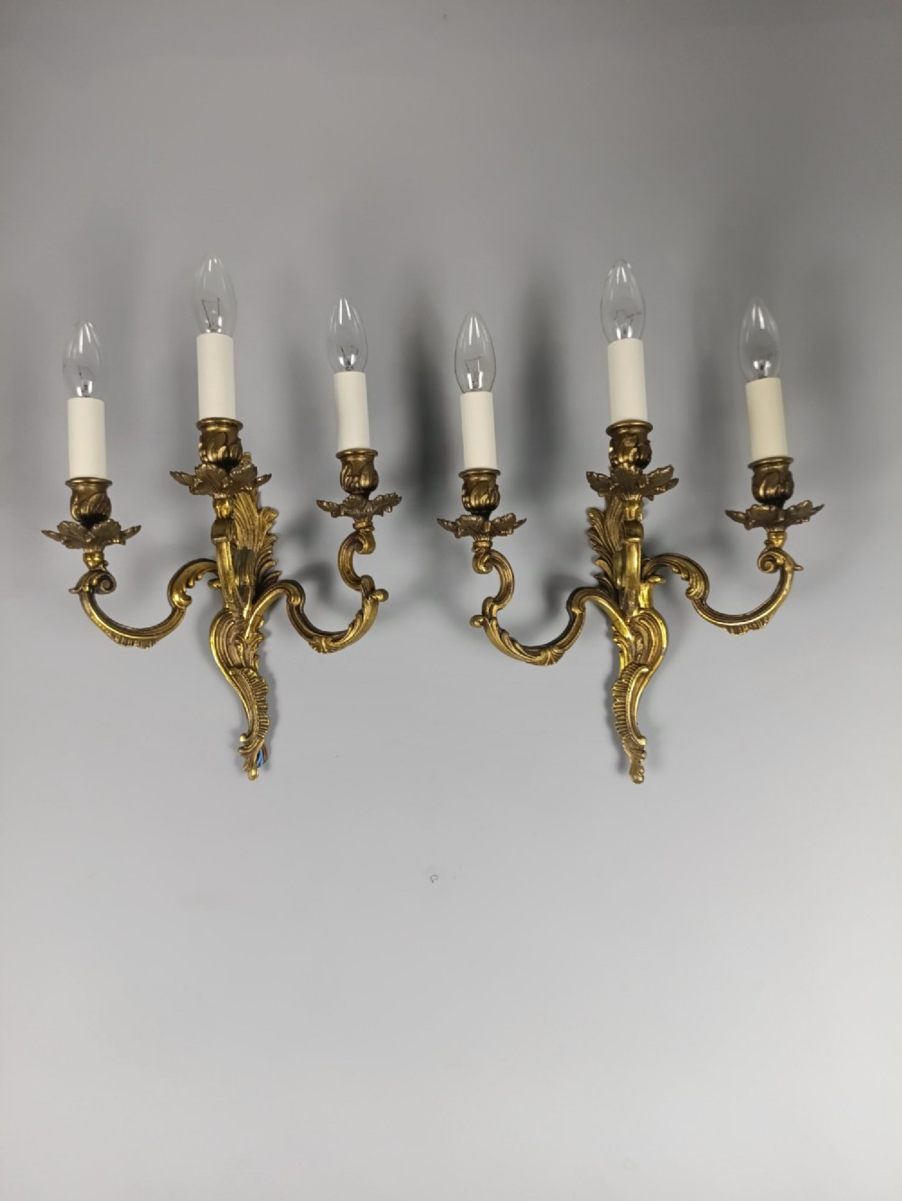 A pair of French Rococo style triple-arm gilded bronze wall lights featuring ornate foliage scrollwork and white candle sleeves. View V
