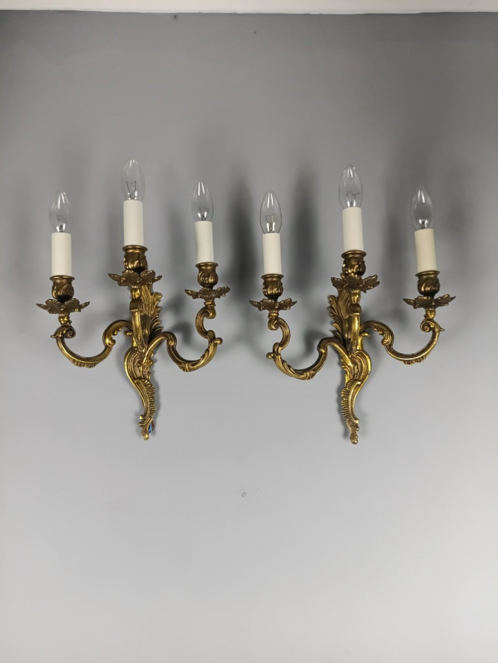 A pair of French Rococo style triple-arm gilded bronze wall lights featuring ornate foliage scrollwork and white candle sleeves. View VI