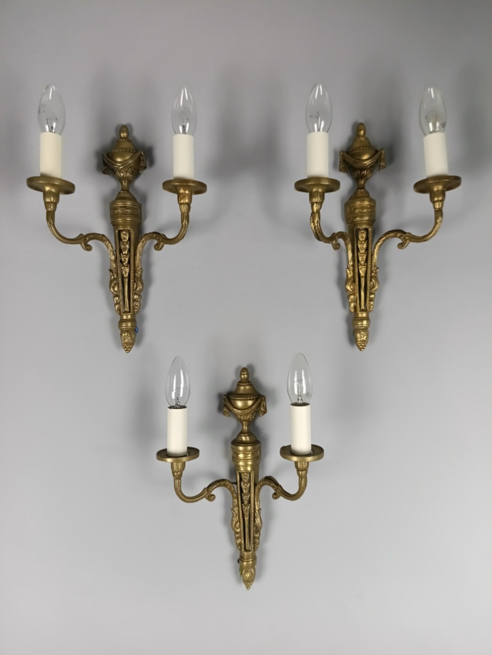 A set of three French Neoclassical gilt brass twin-arm wall sconces featuring urn finials and decorative candle branches.