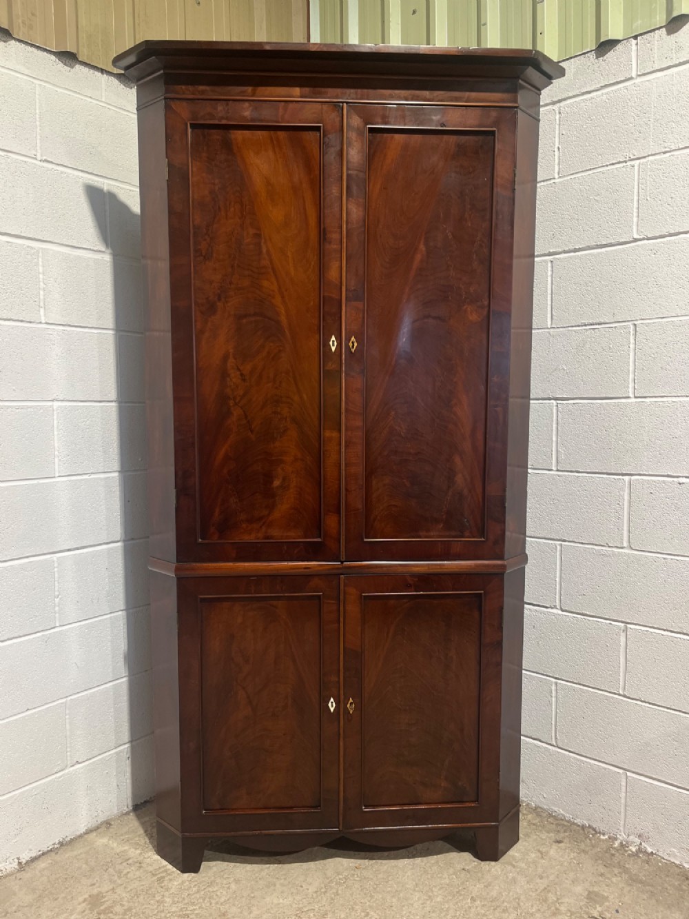 A tall Georgian floor-standing corner cupboard crafted from dark flame mahogany with upper and lower double-panelled doors and brass escutcheons.