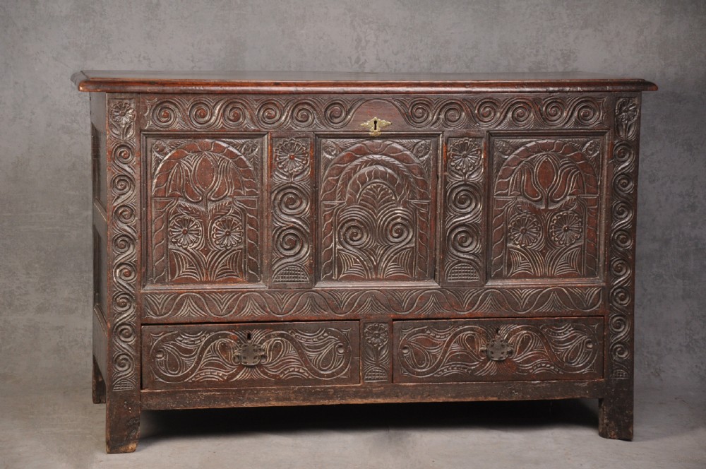 A seventeenth-century carved dark oak Yorkshire coffer featuring three arcaded floral panels, scrollwork friezes, two lower drawers, and brass keyhole escutcheons.