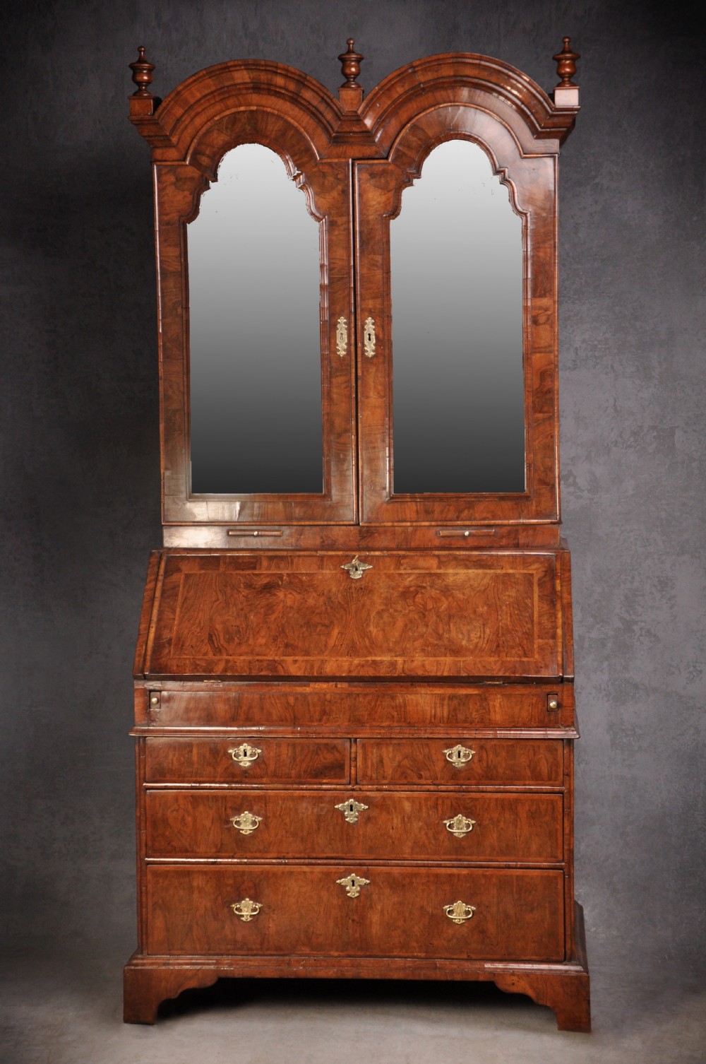 An early 18th-century walnut bureau bookcase with a double-domed top, turned finials, two arched mirrored doors, a slant-front writing slope, and drawers on bracket feet.