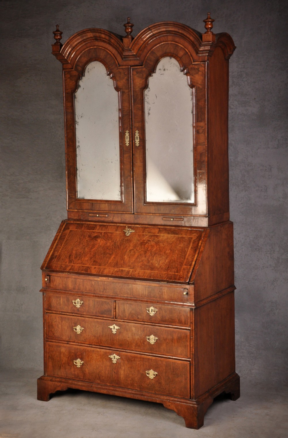 An early 18th-century walnut bureau bookcase with a double-domed top, turned finials, two arched mirrored doors, a slant-front writing slope, and drawers on bracket feet. View II
