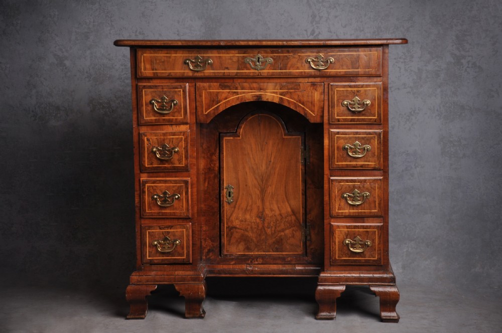 An early 18th-century walnut kneehole desk featuring crossbanded drawer fronts, brass handles, a central recessed cupboard, and bracket feet.