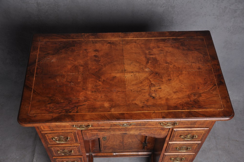 An early 18th-century walnut kneehole desk featuring crossbanded drawer fronts, brass handles, a central recessed cupboard, and bracket feet. View II