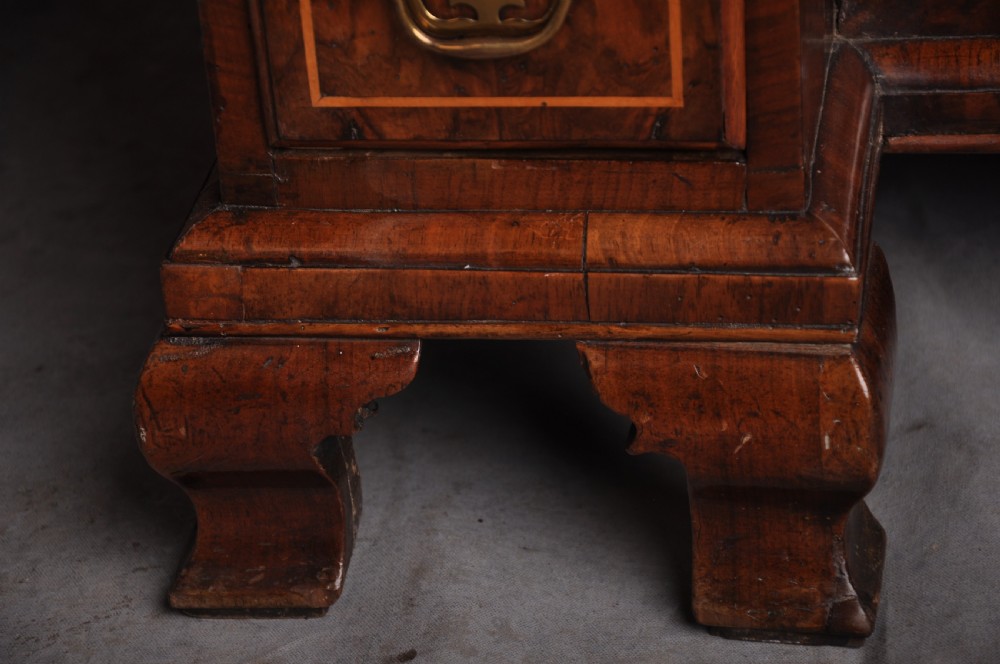 An early 18th-century walnut kneehole desk featuring crossbanded drawer fronts, brass handles, a central recessed cupboard, and bracket feet. View VI