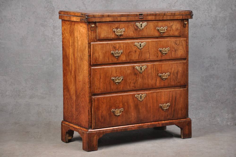 An early eighteenth century walnut bachelor chest featuring a fold-over top, four graduated drawers with brass handles, and bracket feet.