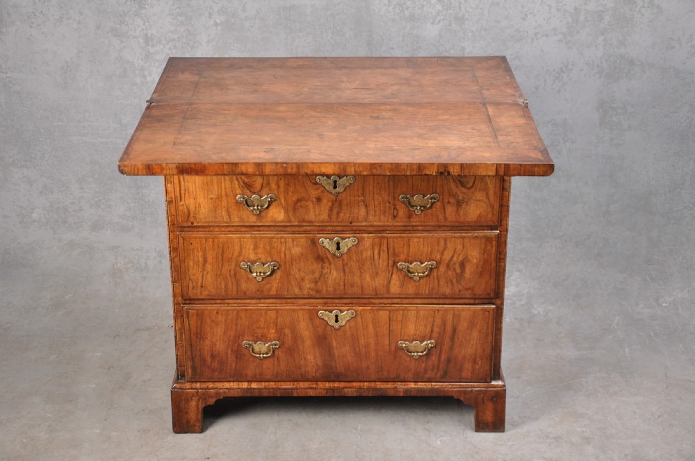 An early eighteenth century walnut bachelor chest featuring a fold-over top, four graduated drawers with brass handles, and bracket feet. View VIII