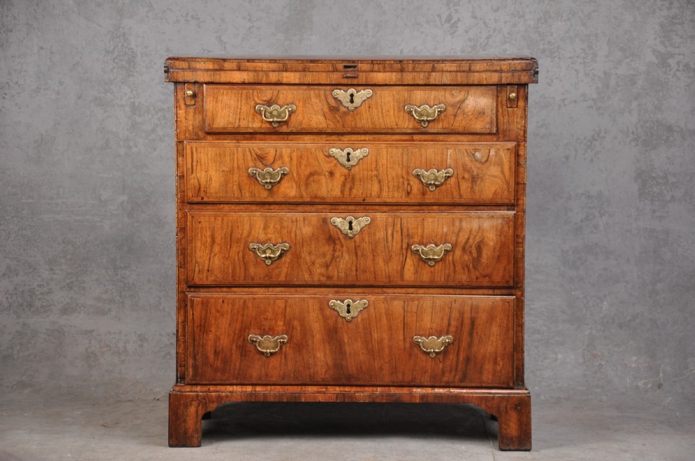 An early eighteenth century walnut bachelor chest featuring a fold-over top, four graduated drawers with brass handles, and bracket feet. View XI