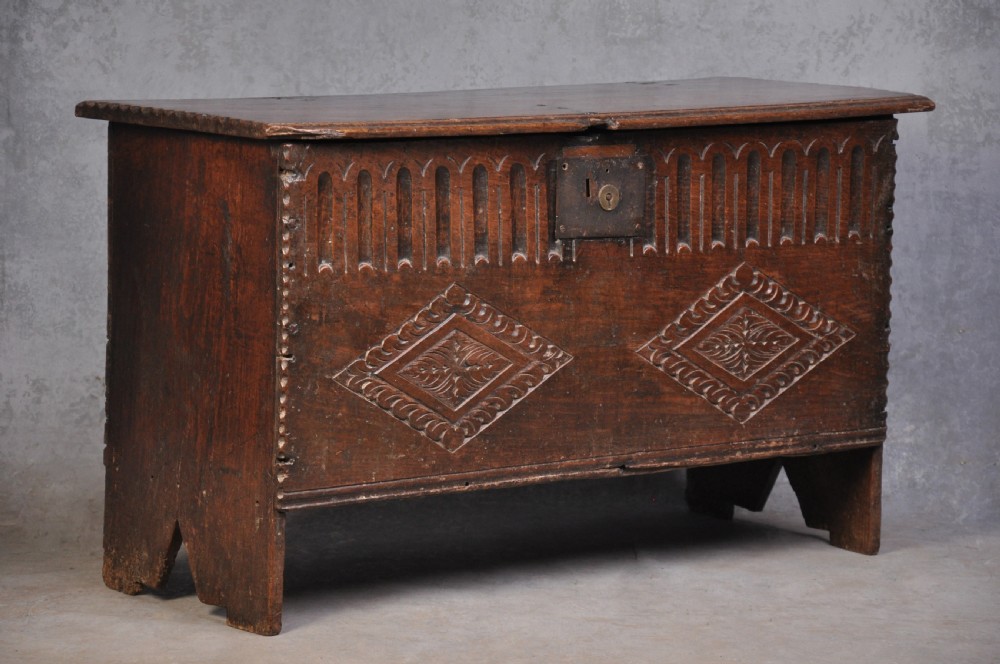 A dark carved oak plank coffer featuring twin carved diamond lozenges, a carved arched top frieze, and a central iron lockplate.