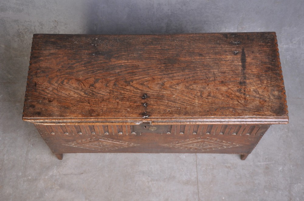 A dark carved oak plank coffer featuring twin carved diamond lozenges, a carved arched top frieze, and a central iron lockplate. View II