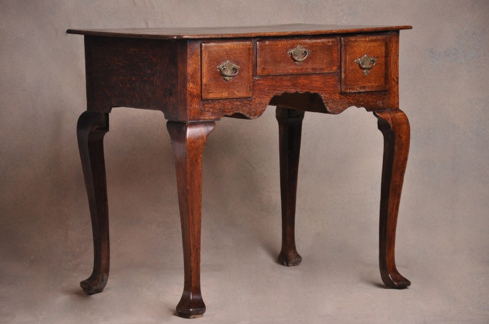 An 18th-century George I oak lowboy featuring three drawers with brass backplate handles, a shaped apron, and four cabriole legs.
