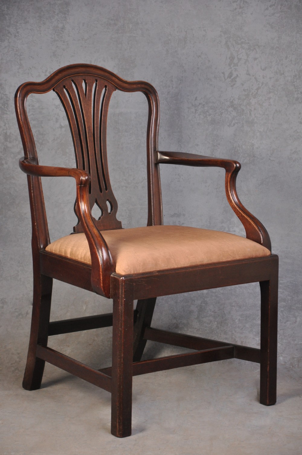 An 18th-century Hepplewhite style mahogany armchair featuring a carved pierced splat, shaped arms, square legs with stretchers, and a beige upholstered seat.