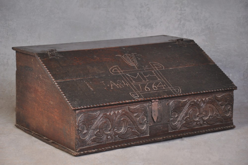 A dark carved oak desk box featuring a sloped lid incised with the date 1664 and initials, resting above foliate carved front panels with iron fittings.