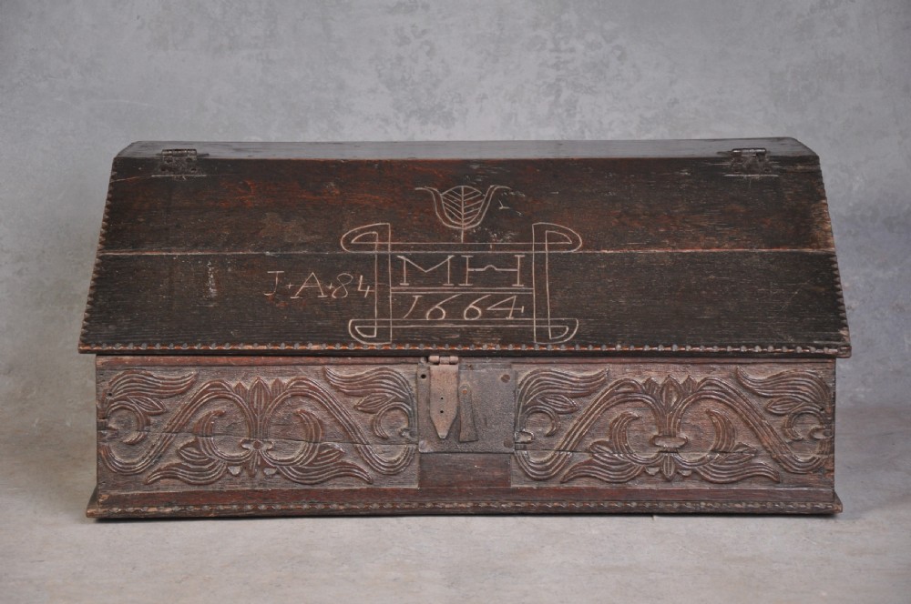 A dark carved oak desk box featuring a sloped lid incised with the date 1664 and initials, resting above foliate carved front panels with iron fittings. View II