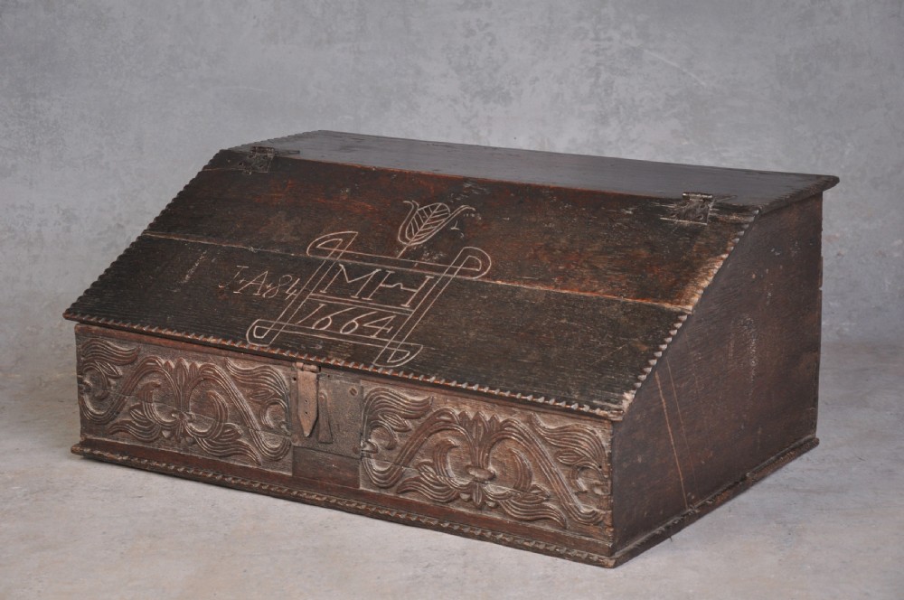 A dark carved oak desk box featuring a sloped lid incised with the date 1664 and initials, resting above foliate carved front panels with iron fittings. View IV