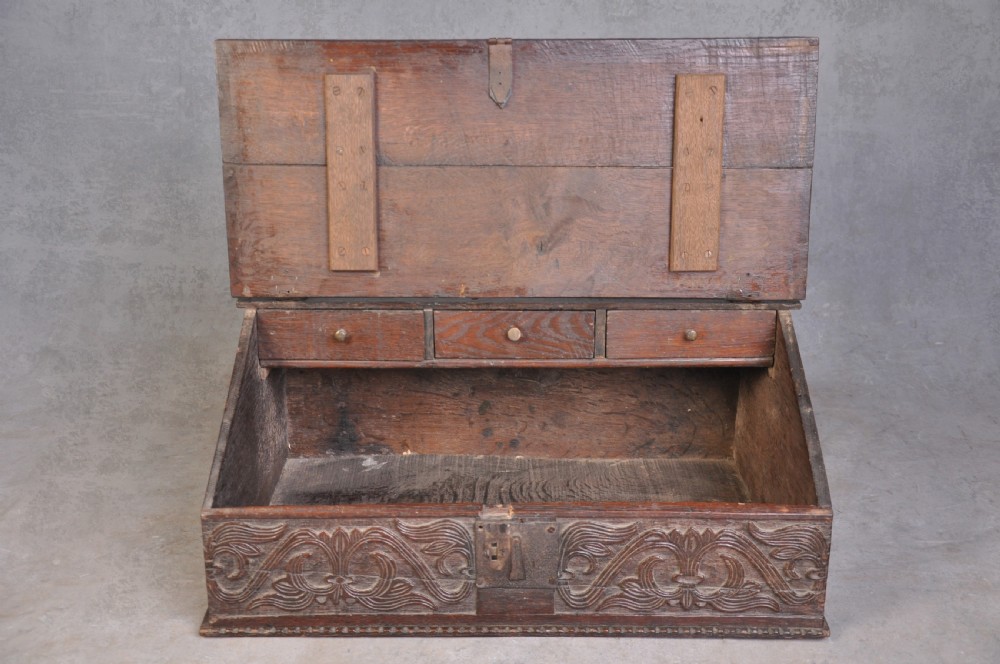 A dark carved oak desk box featuring a sloped lid incised with the date 1664 and initials, resting above foliate carved front panels with iron fittings. View VII